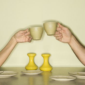 Caucasian mid-adult male and female hands toasting with coffee cups across retro kitchen table setting.