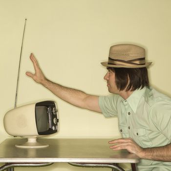 Side view of Caucasian mid-adult man wearing hat sitting at 50's retro dinette set adjusting old television antenna.