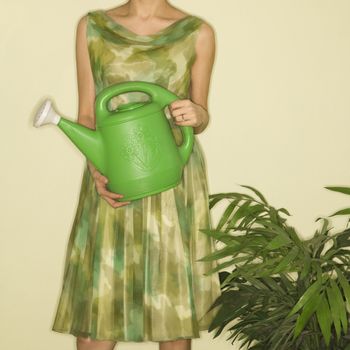 Pretty Caucasian mid-adult woman wearing vintage dress standing next to houseplant holding green watering can.