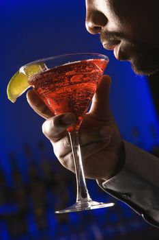 African American man bringing martini up to lips in bar against glowing blue background.