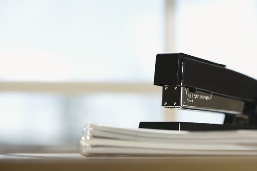 Black stapler on stack of paper with window in background.