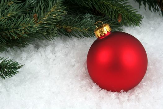 A red Christmas bauble sitting in a bed of snow with an evergreen branch.
