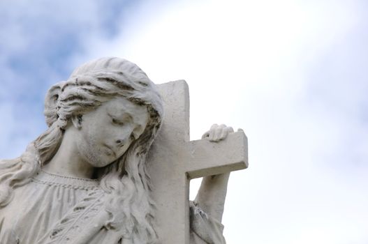 A statue of a angel/girl holding and leaning on a cross with a sorrowful look on face