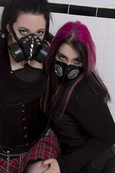 crazy looking teenage girls wearing goth inspired clothes with pink and black hair and gas mask against a white ceramic wall