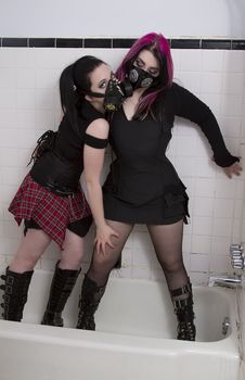 crazy looking teenage girls standing in a tub wearing goth inspired clothes with pink and black hair and gas mask against a white ceramic wall