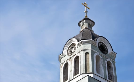 Church bell tower with a clock and a cross against the blue sky.