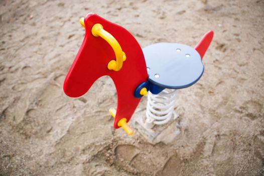 A brightly colored red, blue and yellow rocking horse in a public playground surrounded by sand. - focus on the head with a shallow depth of field.