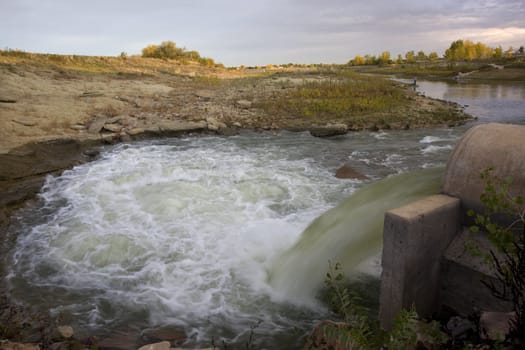 A big pipe supplying water to low level Boyd Lake (Colorado) from another reservoir. There is a distant unrecognizable fisherman figure. October fall colors in cottonwood trees.