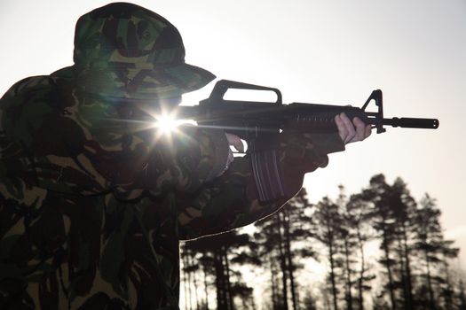 A soldier standing up pointing a rifle foward, covered with camouflage with the Sun shining through.