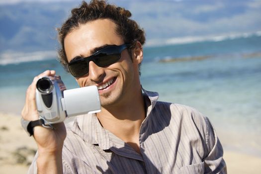 Mid-adult Caucasian man on beach pointing video camera at viewer.