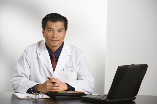 Asian American male doctor sitting at desk with charts and laptop computer looking at viewer.