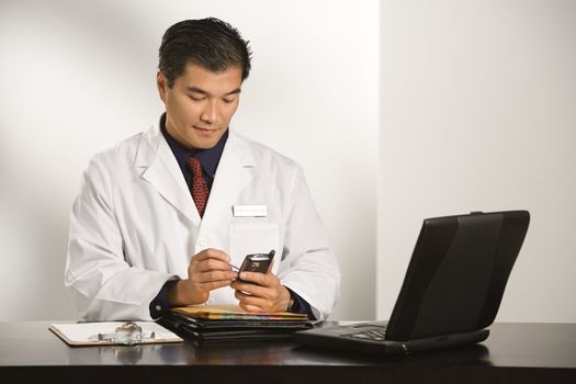 Asian American male doctor sitting at desk with charts and laptop computer using pda.