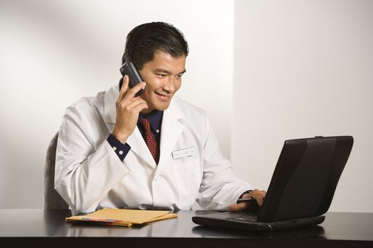 Asian American male doctor sitting at desk with charts and laptop computer talking on cellphone.