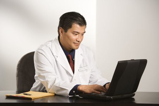 Asian American male doctor sitting at desk with charts typing on laptop computer.