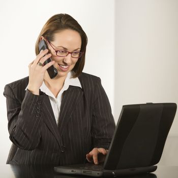 Caucasian businesswoman sitting at desk smiling with laptop computer talking on cellphone.