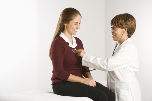 African American middle-aged female doctor examining Caucasian mid-adult female patient with stethoscope.