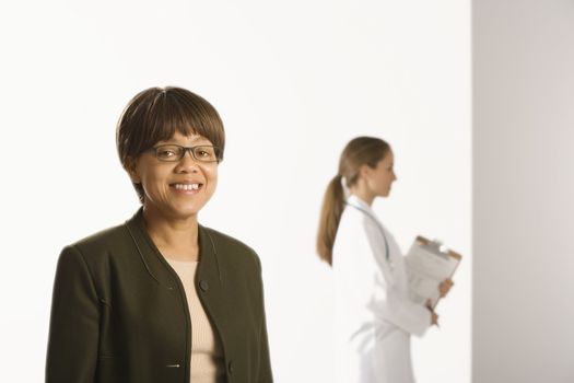 African American middle-aged female patient woman smiling looking at viewer with Caucasian mid-adult female doctor standing in background.