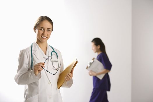 Caucasian mid-adult female doctor smiling and looking at viewer with Asian Chinese mid-adult female physician's assistant walking by in background.
