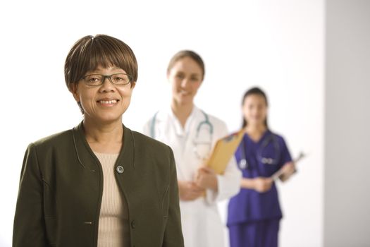 African American middle-aged woman smiling and looking at viewer with Caucasian mid-adult female doctor and Asian Chinese mid-adult female physician's assistant standing in background.