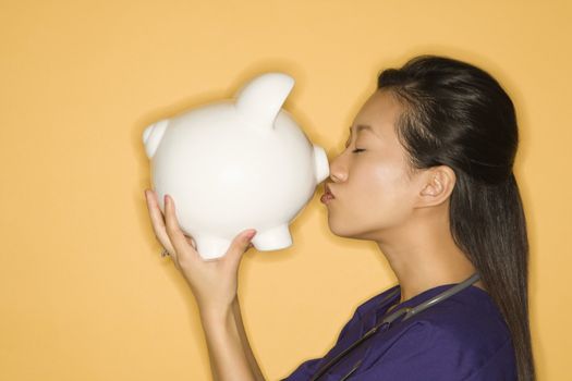 Asian Chinese mid-adult female doctor kissing piggy bank against yellow background.