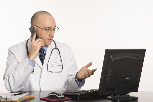 Caucasian mid adult male physician sitting at desk with laptop computer talking on cellphone.