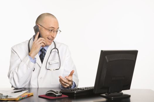 Caucasian mid adult male physician sitting at desk with laptop computer talking on cellphone.
