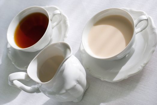 Two cups of tea (one with milk) and porcelain milk jug on the white tablecloth.Shallow depth of field (focus on the edge of the cup from the right).