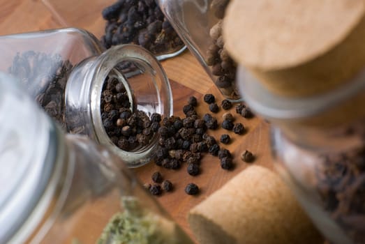 Spilled black pepper seeds from glass jar onto cutting board among other condiments - allspice,juniper etc.