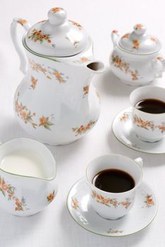 Coffee (or tea) floral pattern service - porcelain tea-kettle, two china cups, milk jug and sugar basin on the white tablecloth. Shallow DOF.