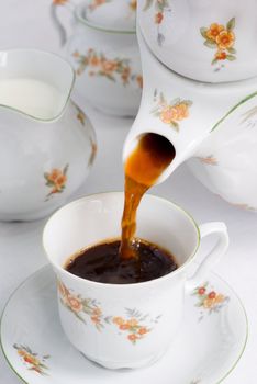 Pouring coffee (or tea) from porcelain tea-kettle into floral pattern porcelain china cup. Milk jug and sugar basin behind. Shallow DOF.