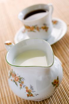 Milk in floral pattern porcelain milk jug on the wooden mat - on the front and cup of coffee behind. Shallow DOF.