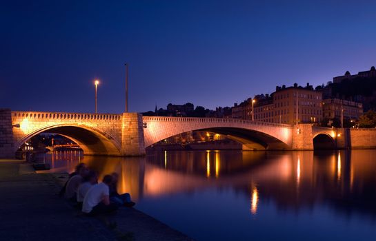 lluminated bridge over the river Saone in the city of Lyon, France, with a group of young people  having a friendly chat by the river