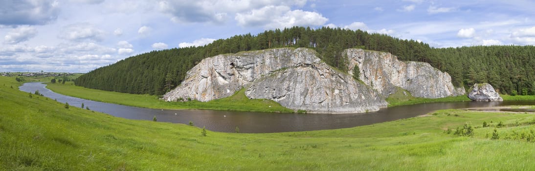 Small river flows between hills. The main object on shot is huge rock with forest. On the left side you can see small village. Shot was taken in june at 15:00.