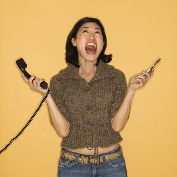 Pretty mid adult Asian woman holding telephone and cellphone with mouth open.