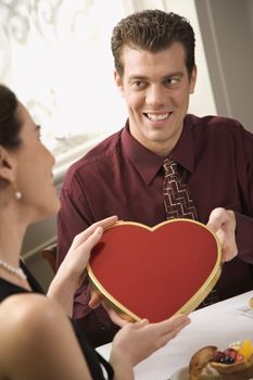 Mid adult Caucasian man giving a heart shaped box of chocolates to surprised woman at restaurant.