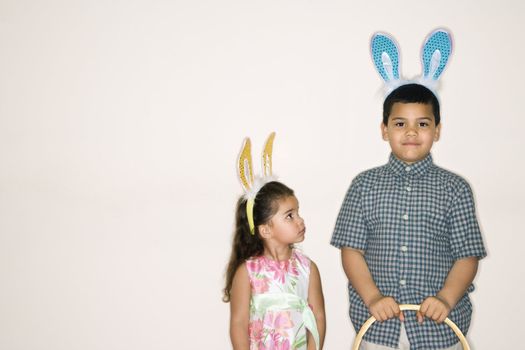 Hispanic girl looking up at Hispanic boy holding Easter basket both wearing bunny ears.