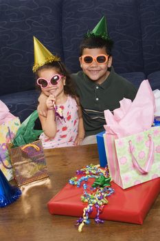 Hispanic brother and sister wearing sunglasses and party hats sitting with presents having a birthday party.