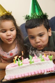 Hispanic girl and boy wearing party hats blowing out candles on birthday cake.