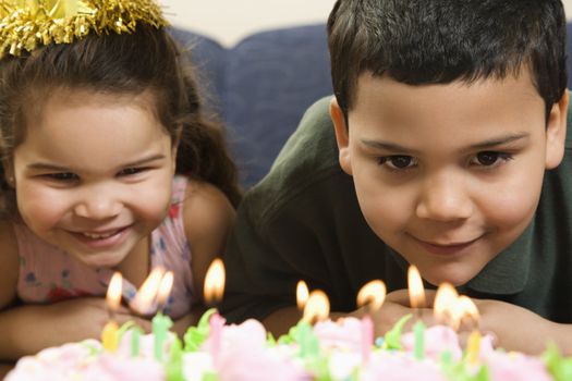 Hispanic girl and boy leaning in close looking at lit candles on birthday cake and smiling.