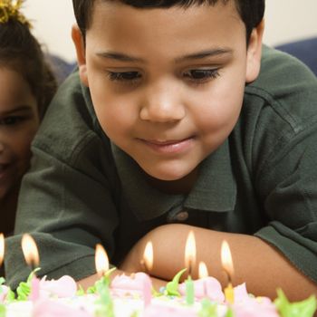Hispanic boy looking down wishfully at lit candles of birthday cake with girl peeking in over his shoulder.