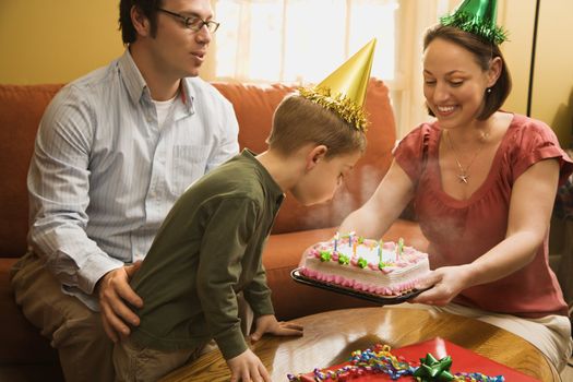 Caucasian boy in party hat blowing out candles on birthday cake with family watching.