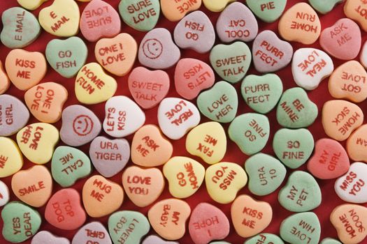 Large group of colorful candy hearts with sayings on them arranged on red background.
