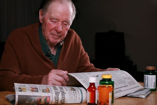 Elderly man reading  news of stock market problems, with medications, money and checkbook on table.