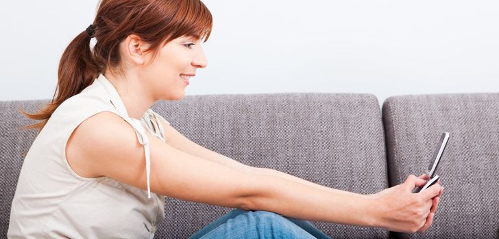 Beautiful woman seated on sofa holding a cellphone and sending a sms