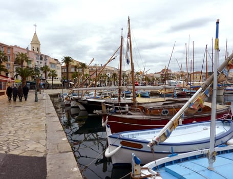 View of Sanary-sur-mer harbor with its colored boats and people walking on the side with a church, palm trees and buildings, France, by cloudy weather