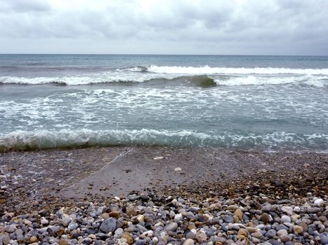 Mediterranean sea and its waves at a beach with lots of pebbles and small brown and grey stone, by cloudy weather
