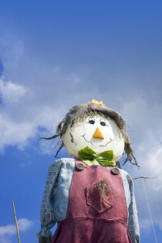 A smiling hand made scarecow stuffed with straw and wearing a red pair of dungarees with a blue checkered shirt , a green felt bow tie and a hesian hat. Set at a low angle against a blue sky background.