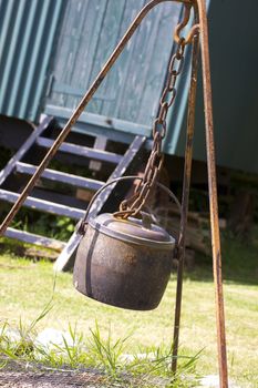 A rusty metal cauldron hanging over an open fire space within a rusty metal frame, hook and chain, set in front of the wooden steps of a green painted tin constructed shepherds hut. Located in rural Dorset, England.