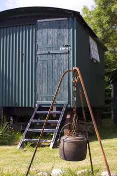 A metal cauldron hanging over an open fire space within a rusty metal frame and hook, set in front of a green painted tin constructed shepherds hut. Located in rural Dorset, England.