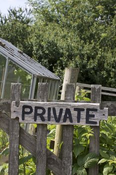 A wooden garden gate with a hand painted sign with the word private nailed to its front. The gate is the entrance to a vegetable garden and allotment located in rural Devonshire countryside, England. A greenhouse is visible to the background.